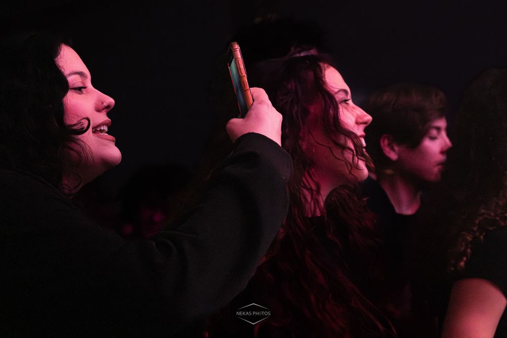 Three young women at a concert, one holding a smartphone up to record, bathed in pink stage lighting.