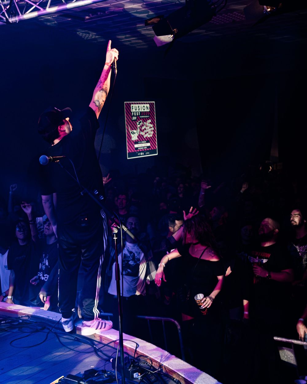 Performer on stage raises fist at dark concert venue with crowd and neon poster backdrop.