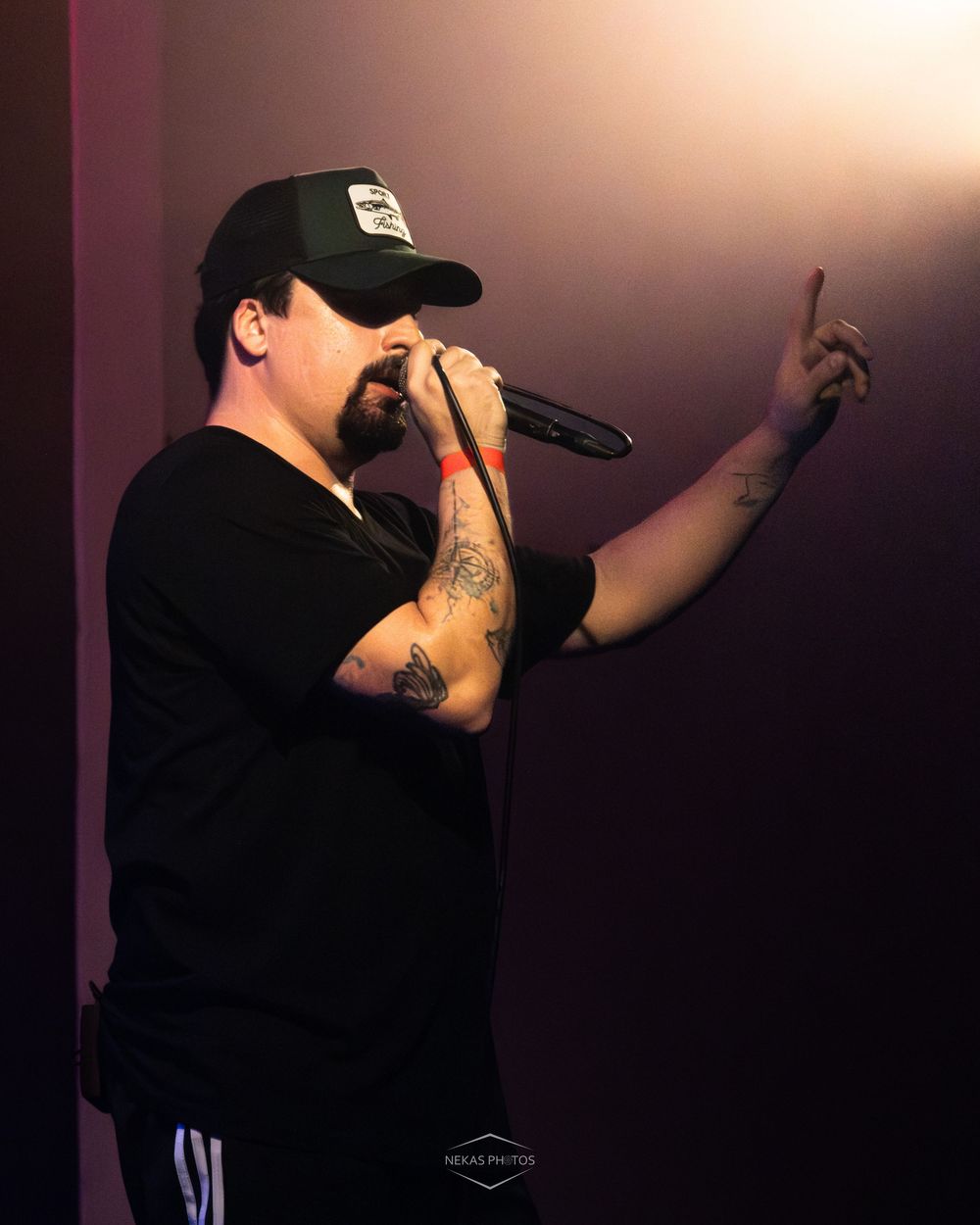Tattooed male rapper performing on stage with microphone, wearing black cap and shirt, pointing finger upward under purple stage lighting.
