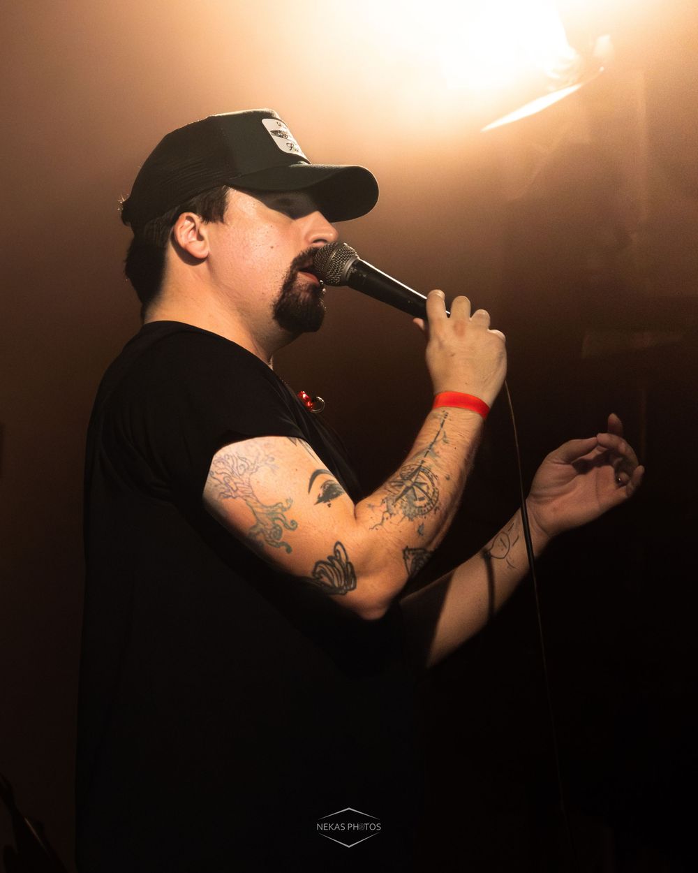 Tattooed male rapper performing on stage with microphone, wearing black cap and t-shirt under warm stage lighting.
