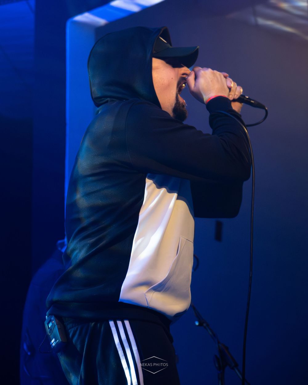 Male hip-hop artist performing on stage in black hoodie and Adidas pants, singing into microphone under blue lights.
