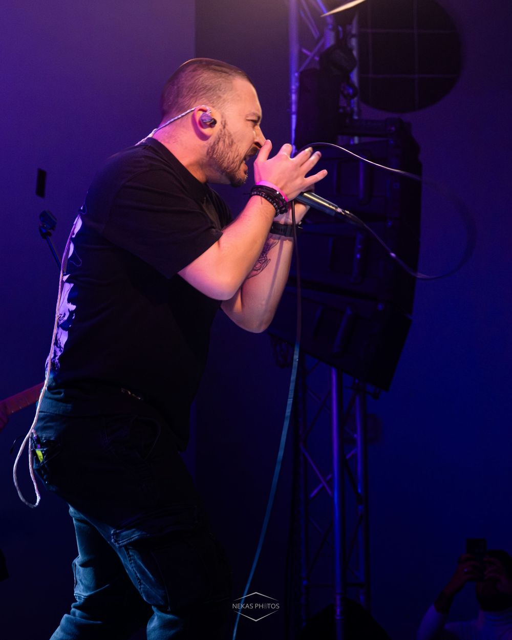 A bearded male vocalist performs passionately on stage under purple lighting, singing into a microphone.