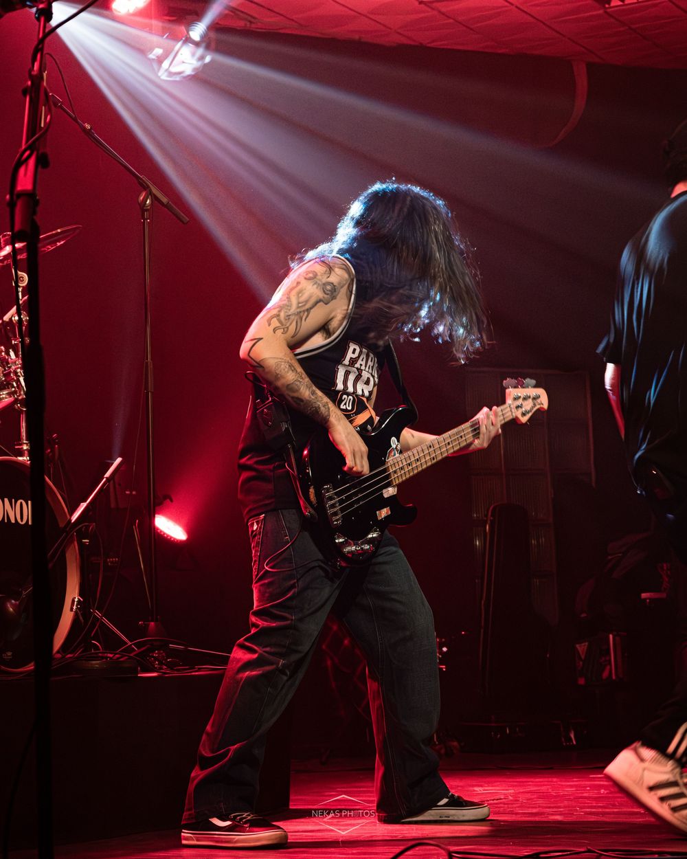 Tattooed bassist with long hair playing electric guitar on a red-lit stage during a live rock concert performance.