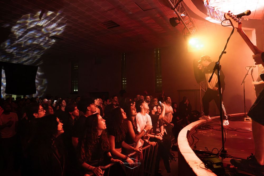 Crowd of concert fans watching a live music performance under colorful stage lights in a large indoor venue.