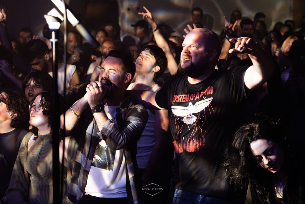 Two male singers performing on stage at a crowded rock concert at night, one wearing a Parkway Drive band t-shirt.