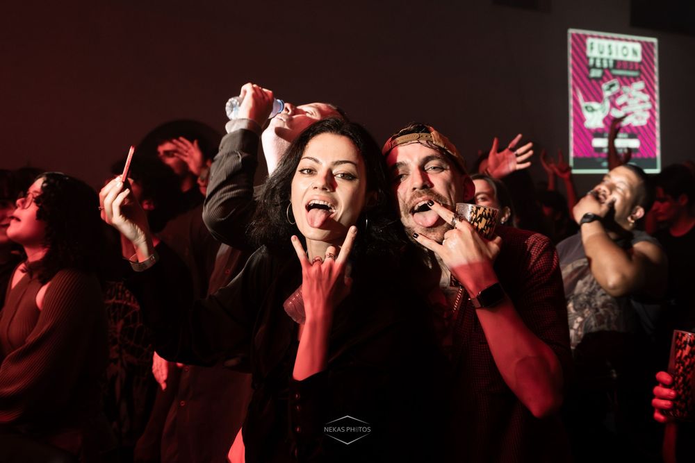 Crowd of rock fans making devil horns gestures at a dark concert venue with red lighting.