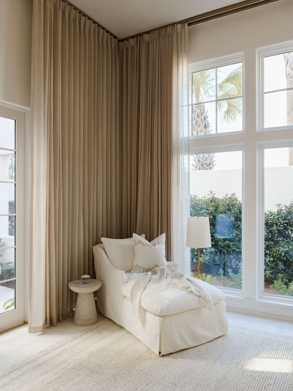Serene reading nook with cream chaise lounge, floor-length beige curtains, and large windows overlooking lush greenery.