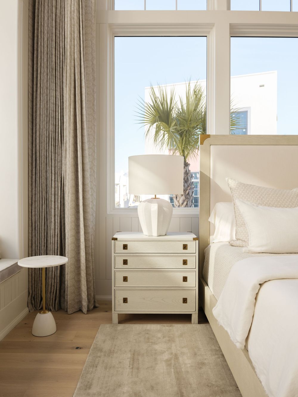 Elegant neutral bedroom with cream nightstand, white lamp, linen drapes, and palm tree view through large windows.