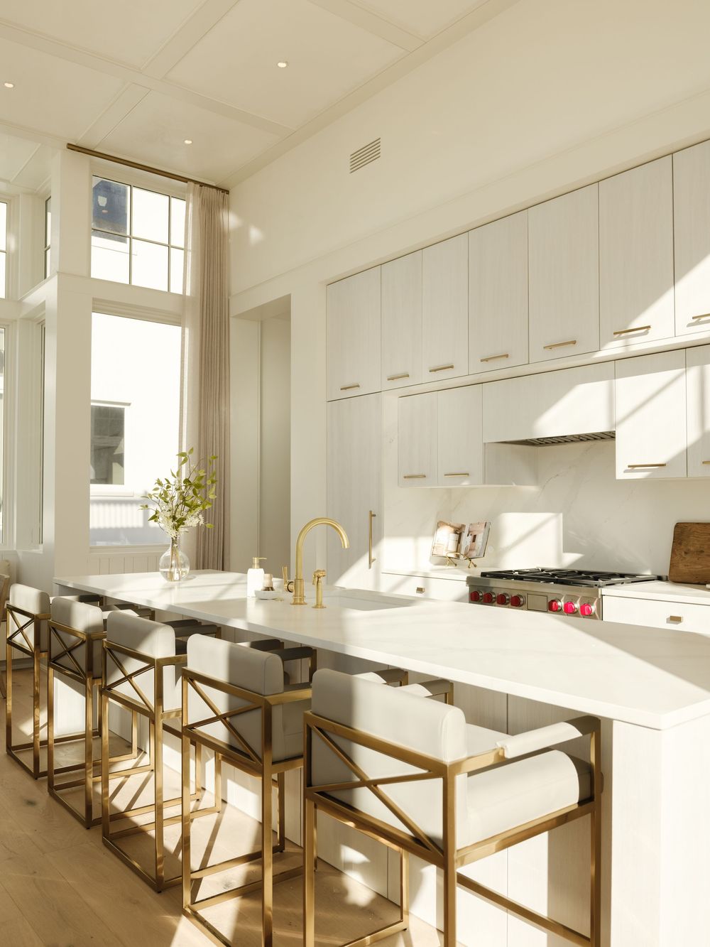 Bright white kitchen with gold bar stools, marble island, brass faucet, and floor-to-ceiling windows flooding the space with natural light.