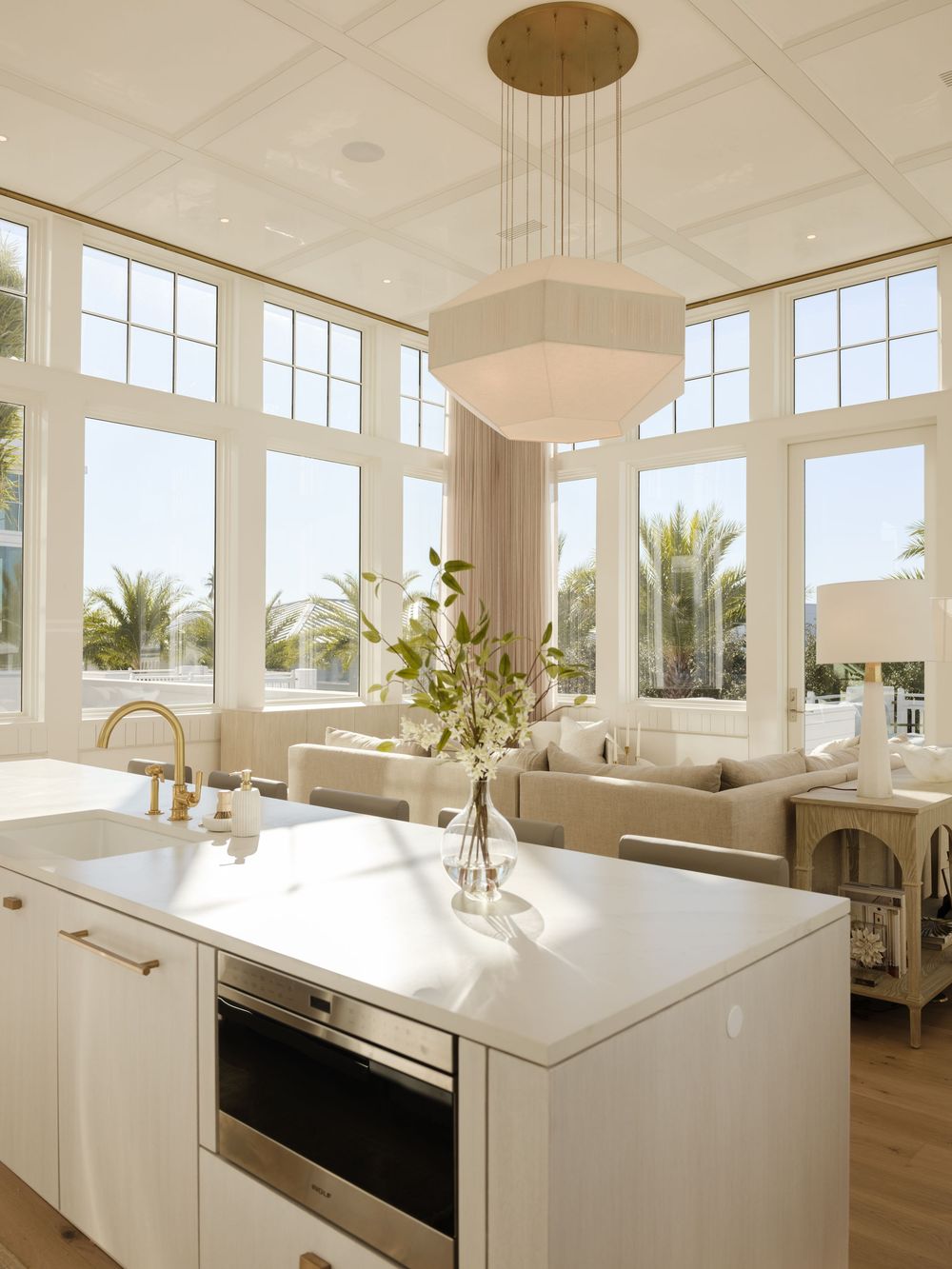 Bright open-plan kitchen with white island, gold fixtures, pendant light, and floor-to-ceiling windows overlooking palm trees.