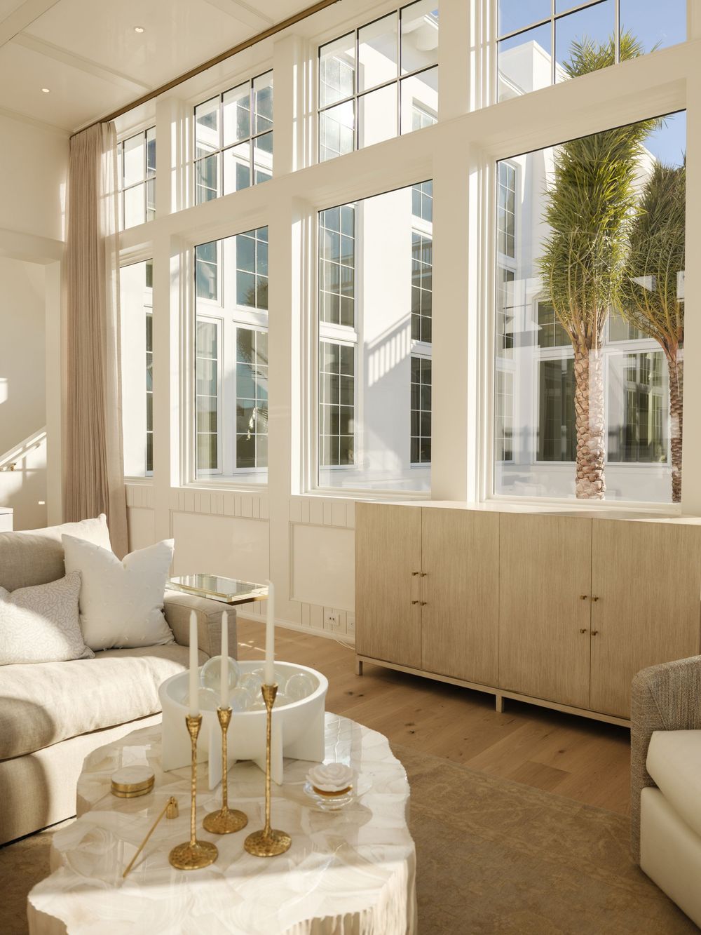 Bright modern living room with floor-to-ceiling windows, neutral tones, marble coffee table, gold accents, and light wood cabinet.