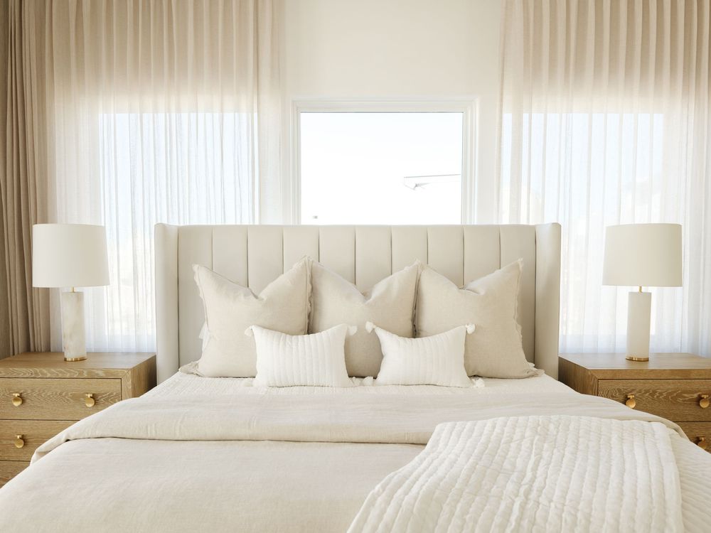 Elegant neutral bedroom with beige upholstered headboard, layered cream pillows, matching nightstands, and sheer white curtains.