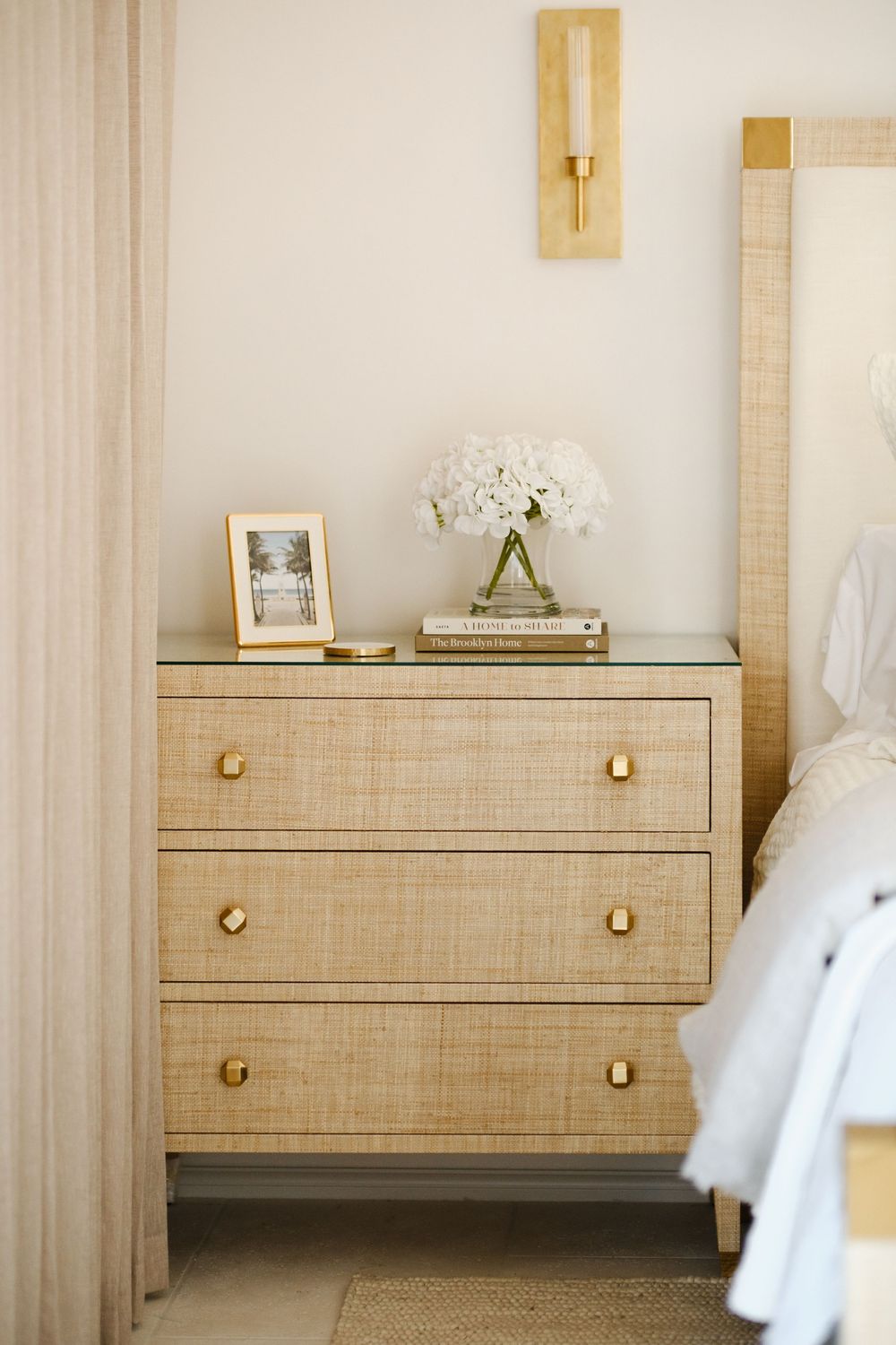Elegant rattan nightstand with gold hardware topped with white flowers, framed photo, and books in a neutral bedroom.