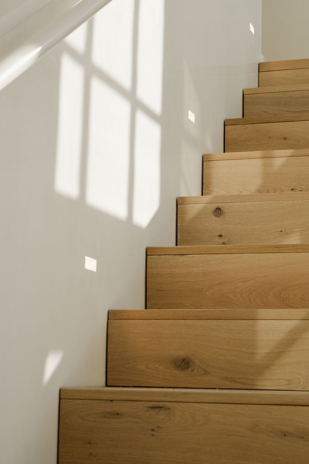 Natural oak wood staircase with white walls and soft window light shadows in a modern minimalist home interior.