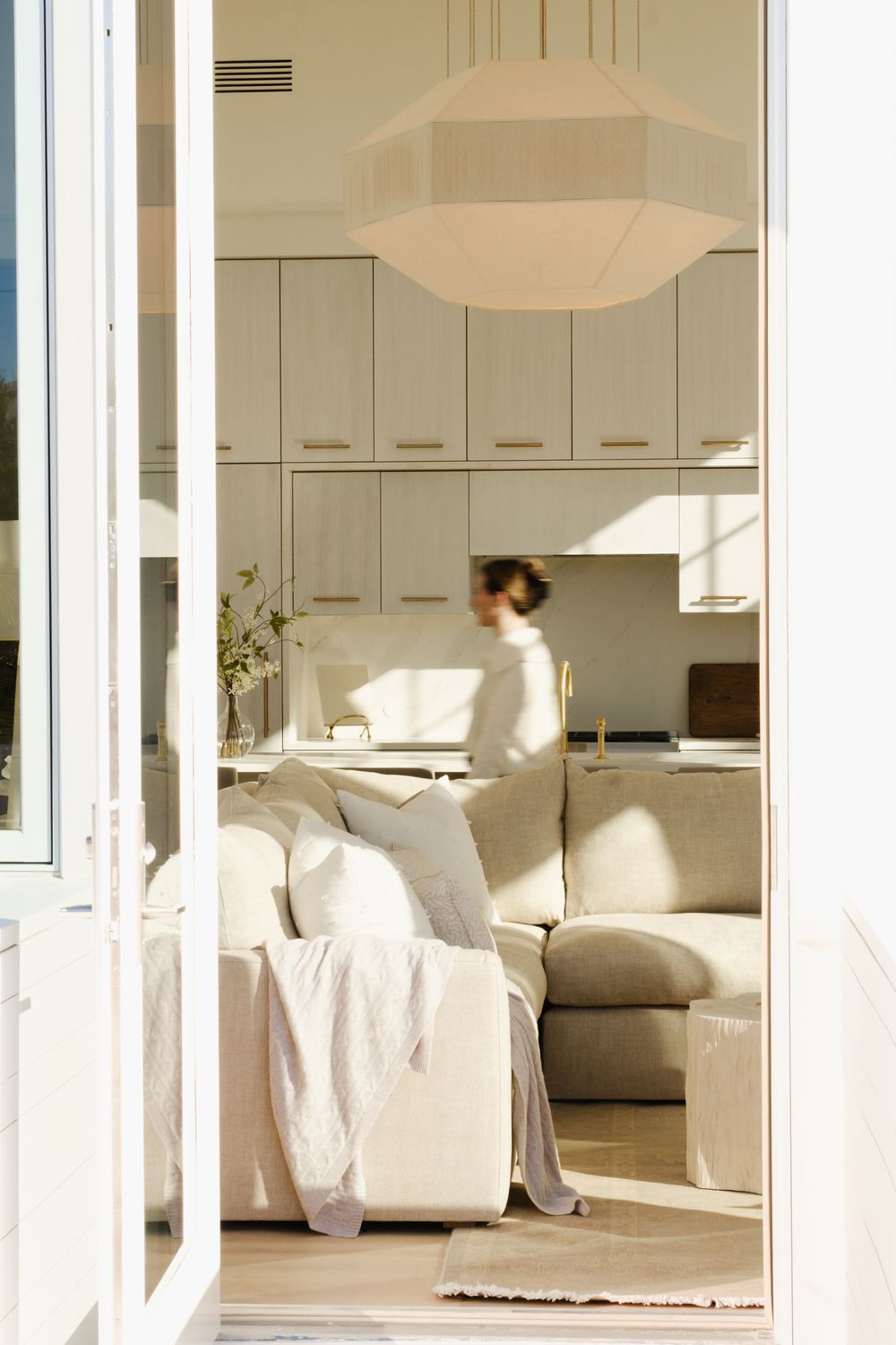 Sunlit open-plan living space with cream sectional sofa, geometric pendant light, and white cabinetry visible through French doors.
