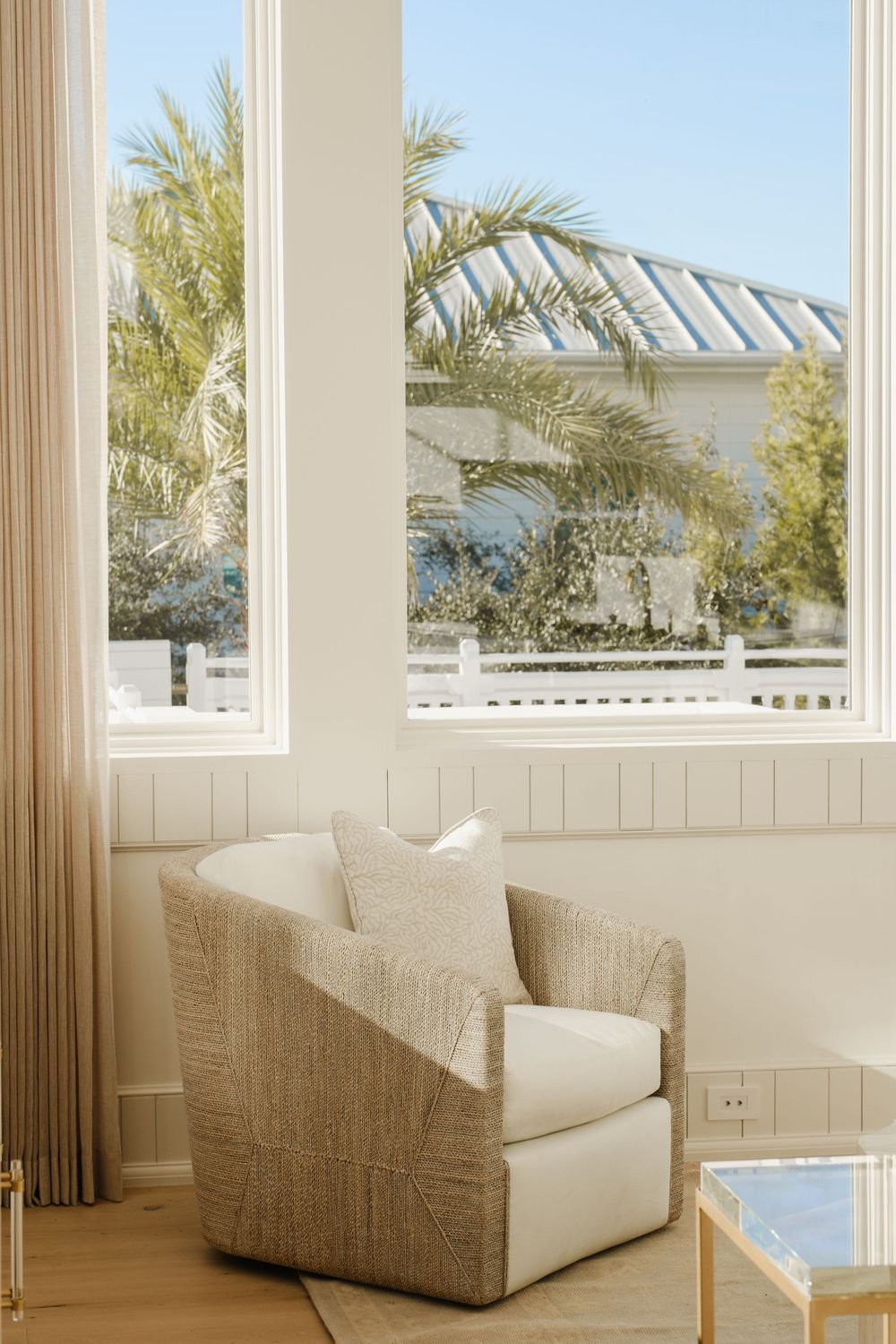 Cozy neutral reading nook with woven swivel chair, cream cushion, and large windows overlooking tropical palm trees and blue sky.