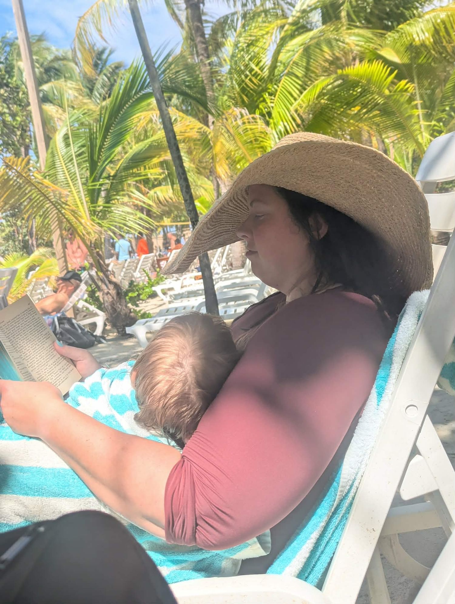 Woman in straw hat breastfeeding baby on lounge chair at tropical beach resort with palm trees.