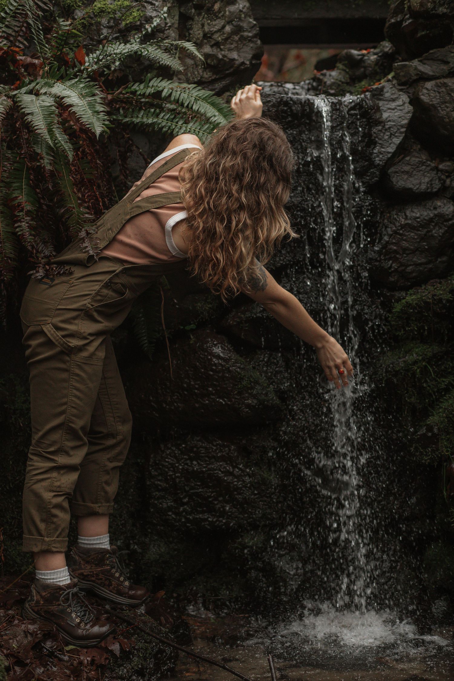 young woman wearing dark green overalls reaching her hand out to touch small waterfall at interlaken park in seattle