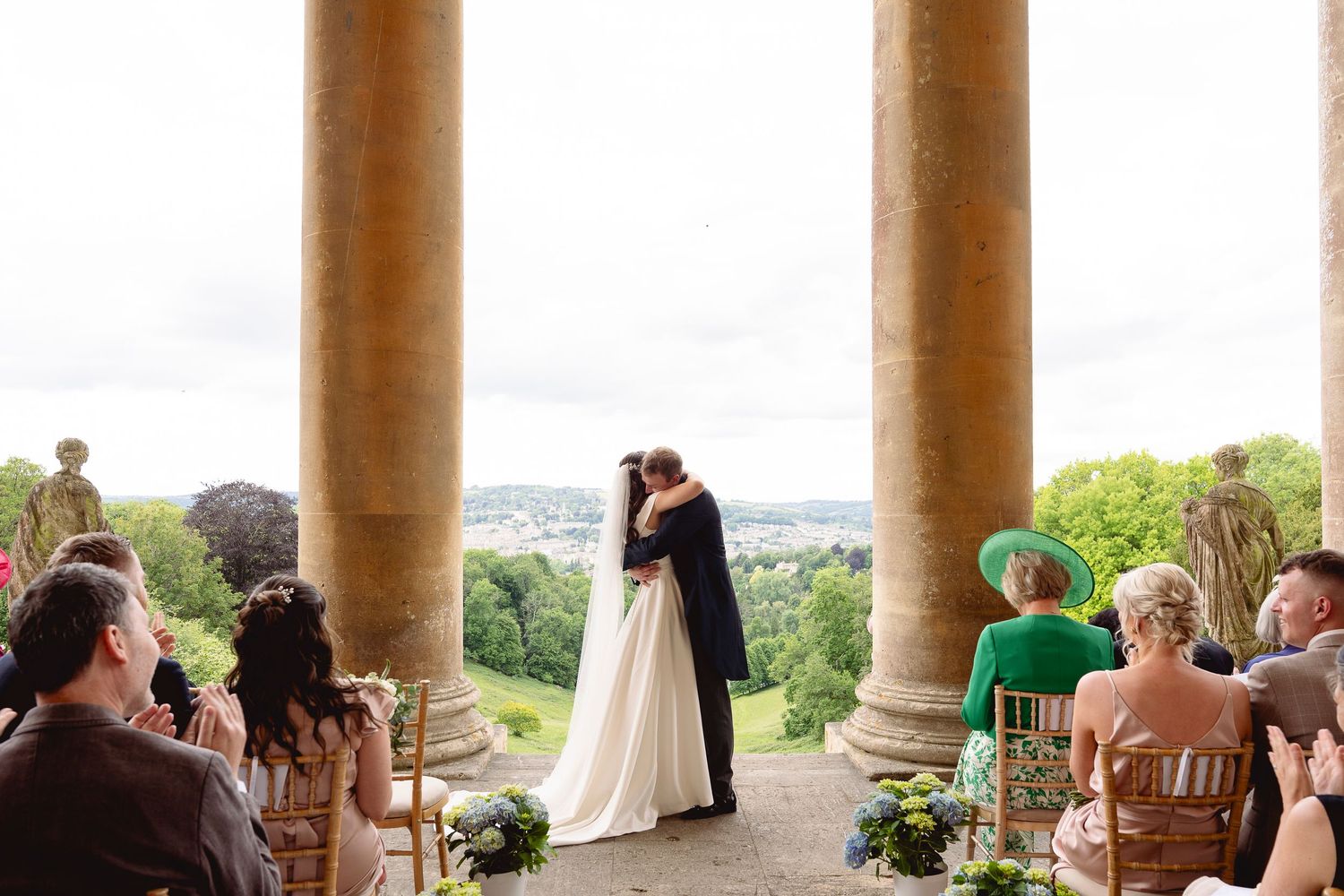 Bride and groom share first kiss at outdoor wedding ceremony with stone columns and scenic countryside views.