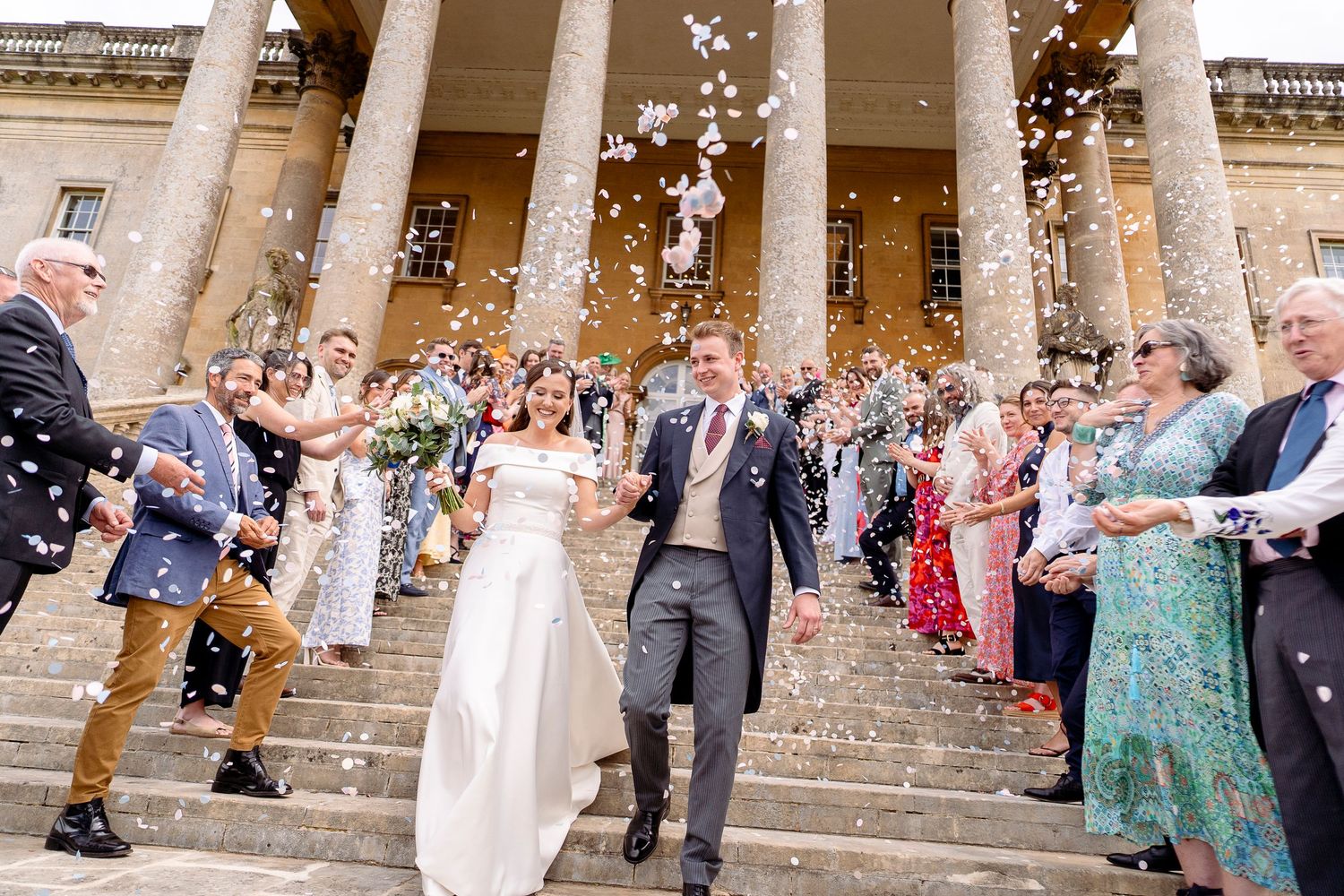 Bride and groom walk through confetti shower on grand columned steps surrounded by cheering wedding guests.