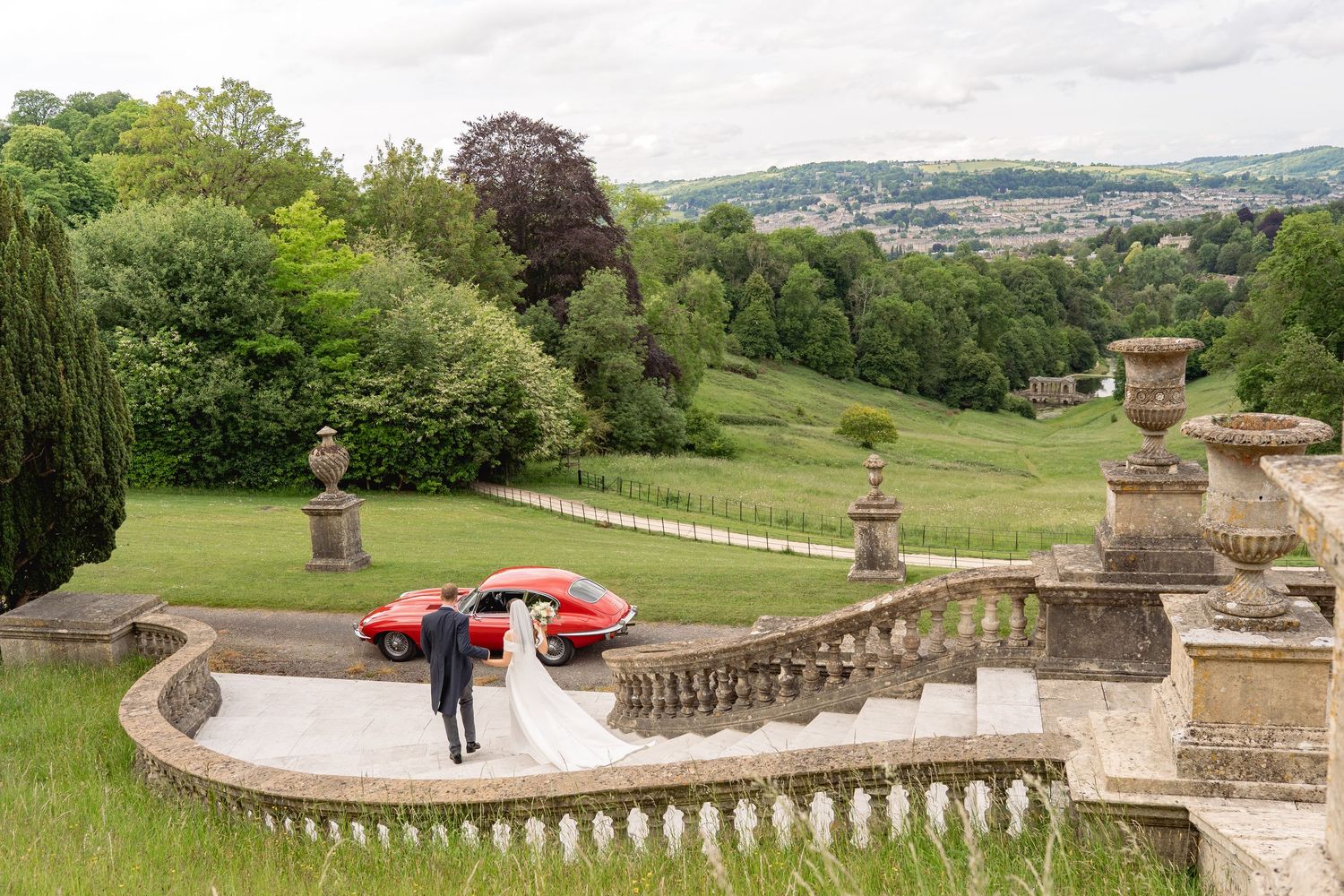 Bride and groom pose by a red sports car on stone terrace of elegant country estate with rolling green landscape.