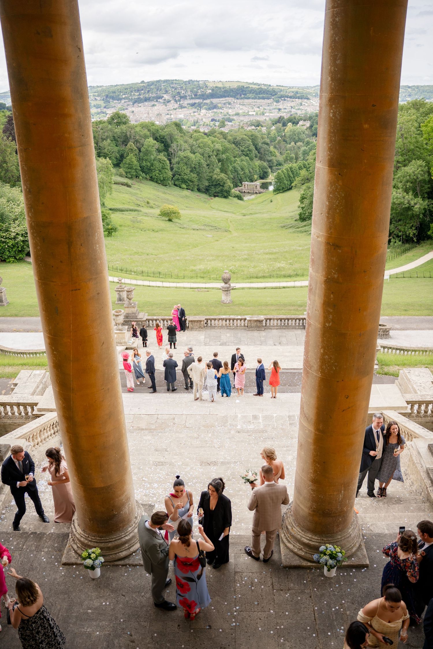 Wedding guests gather on grand stone steps flanked by tall columns, overlooking lush green hills and a distant cityscape.