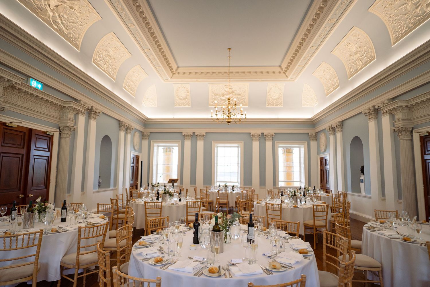 Elegant wedding reception hall with ornate plasterwork ceilings, gold chiavari chairs, and round tables with white linens.