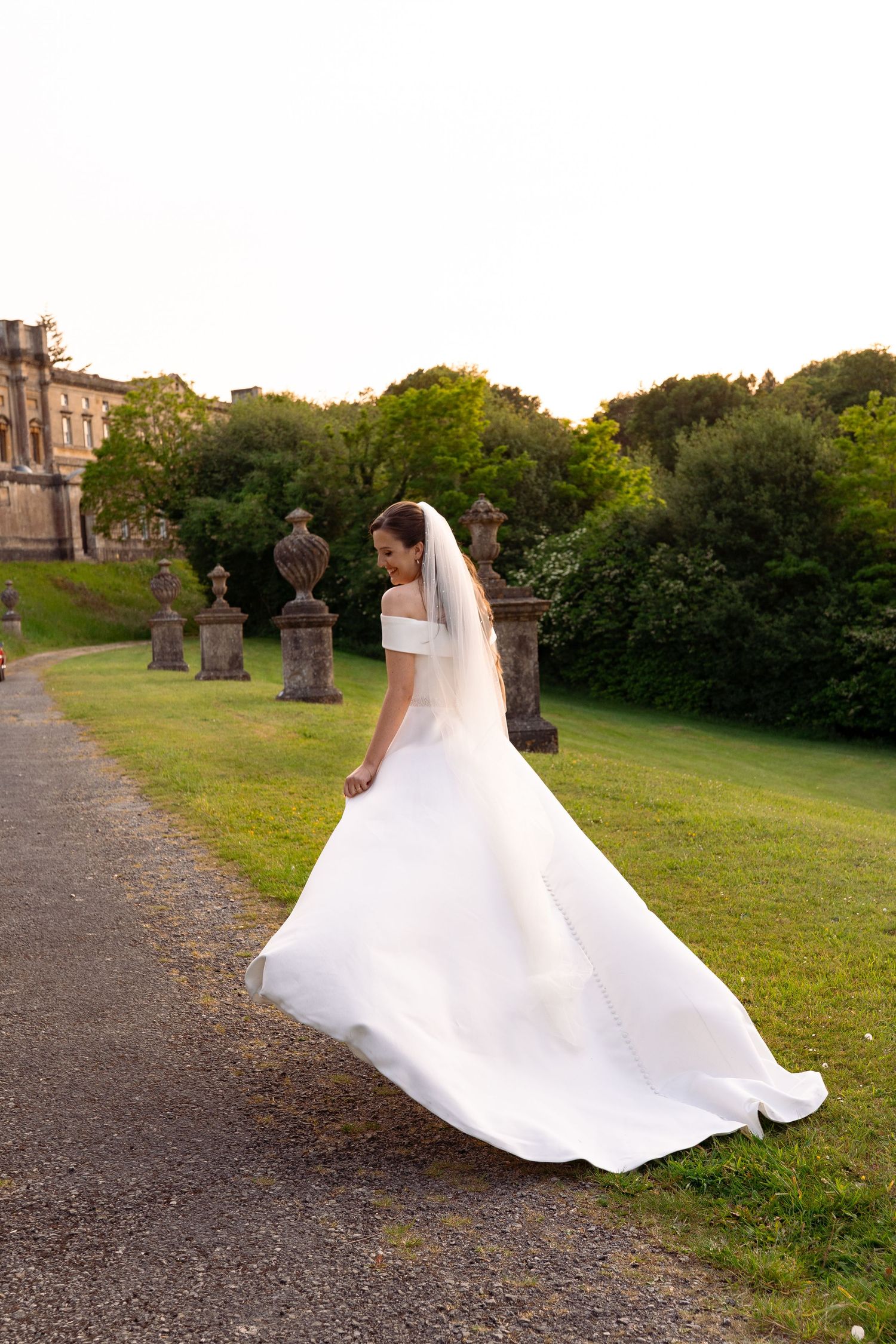 Bride in elegant white off-shoulder gown with veil walks on garden path at historic estate during golden hour.