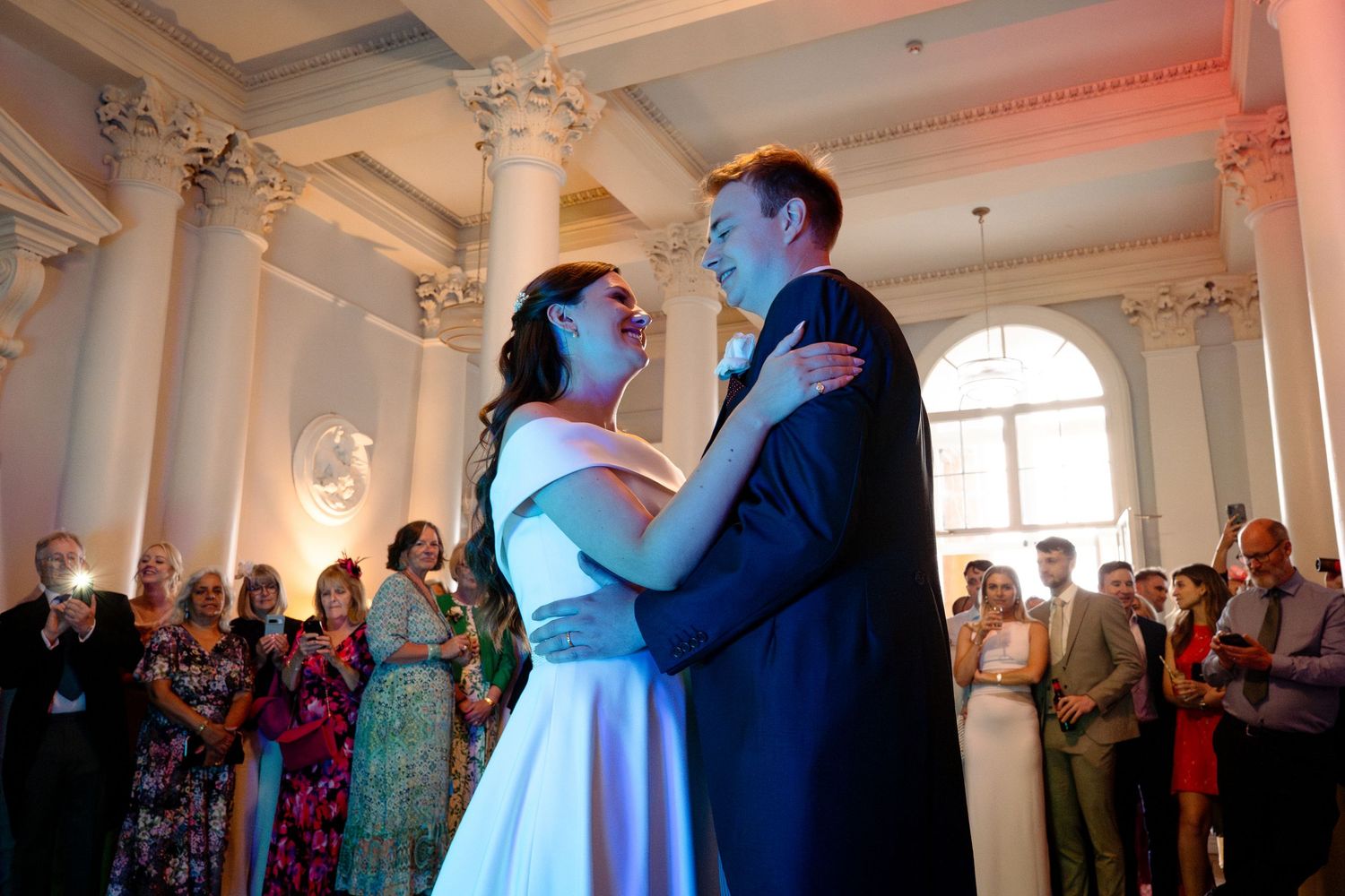 Bride and groom share first dance at elegant wedding reception with guests watching in grand hall.