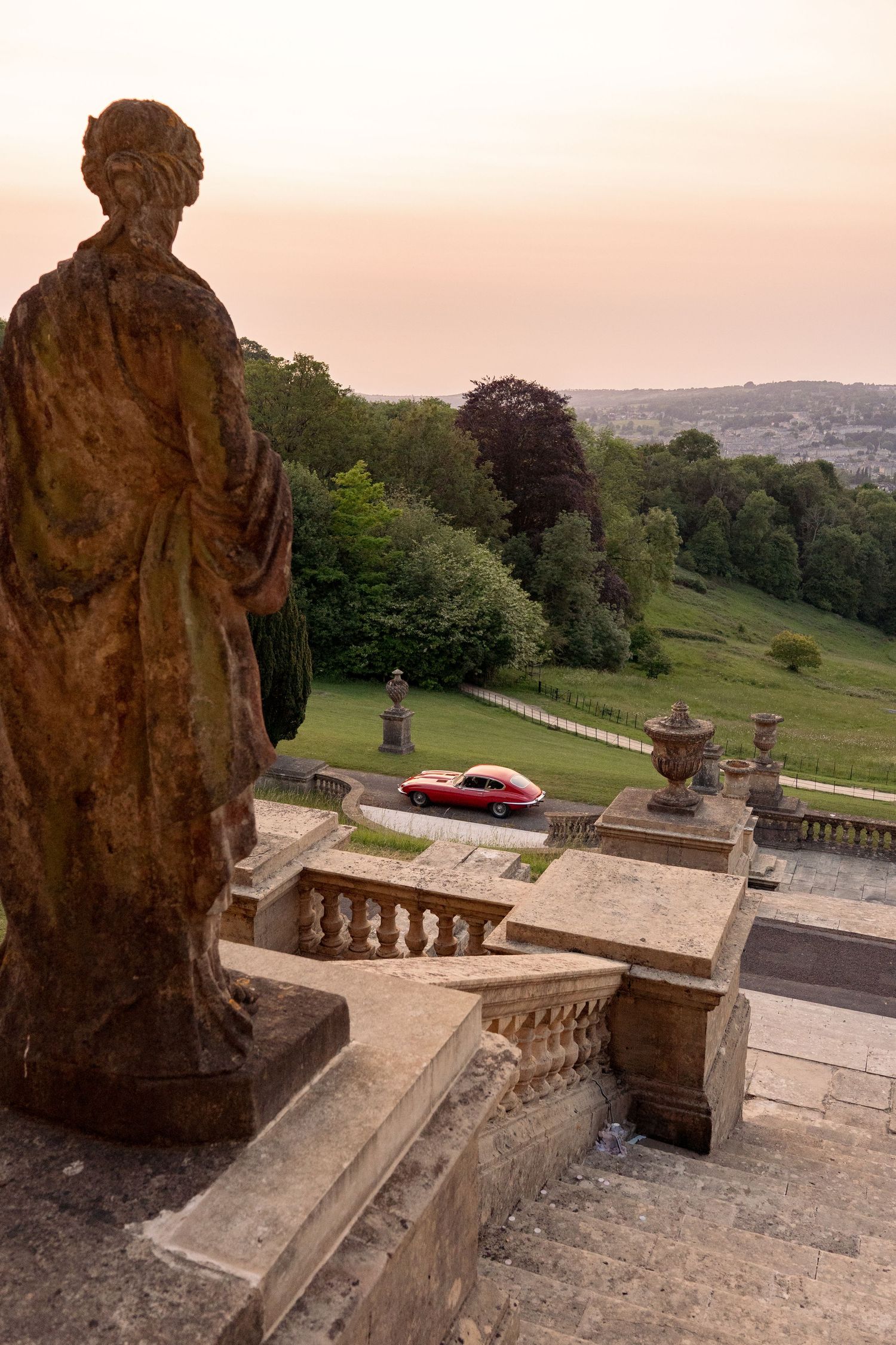 Stone statue overlooks grand staircase with red vintage car driving past lush green landscape at golden hour sunset.