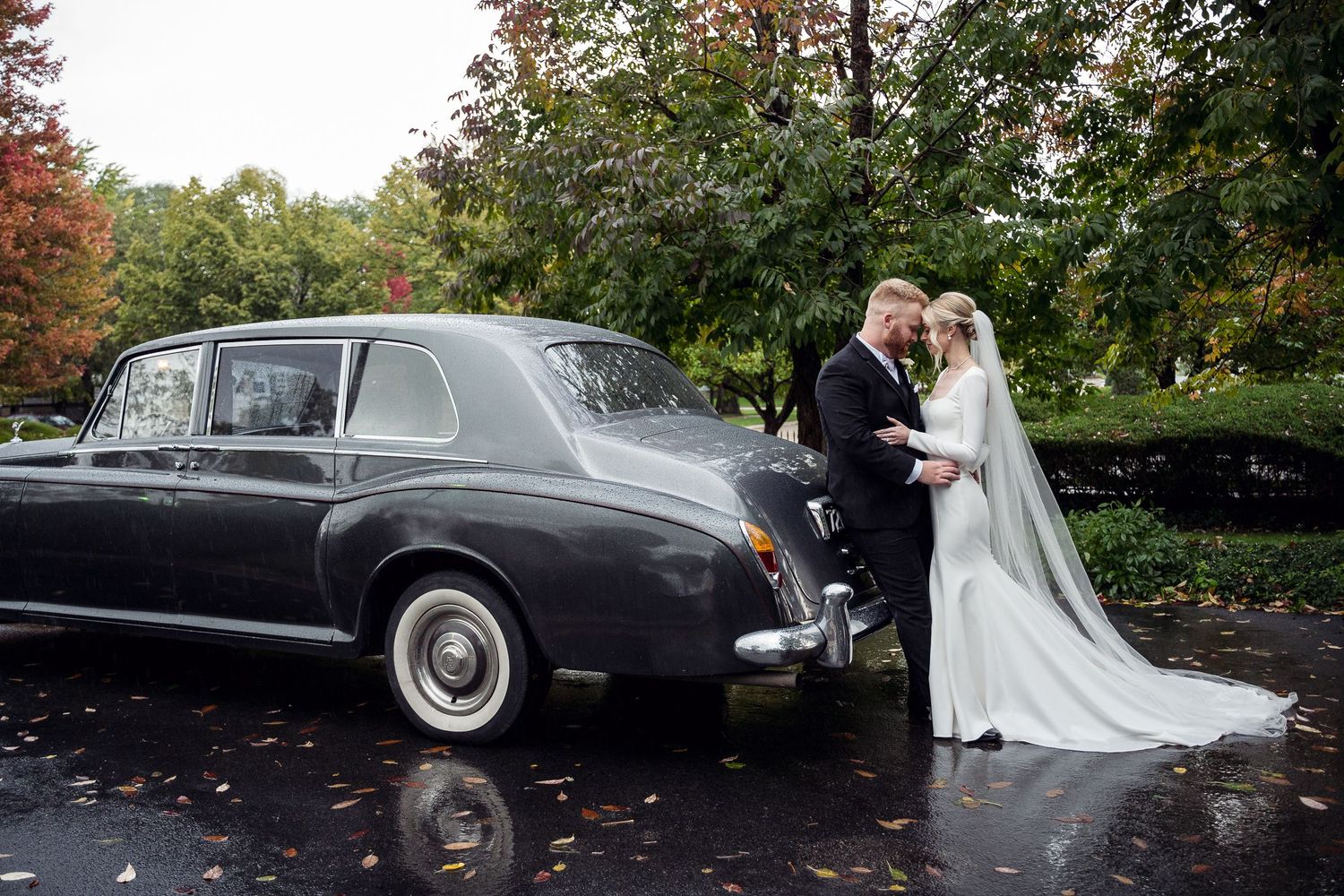 Bride and groom share a romantic moment beside a classic gray Rolls-Royce on a rainy autumn wedding day.