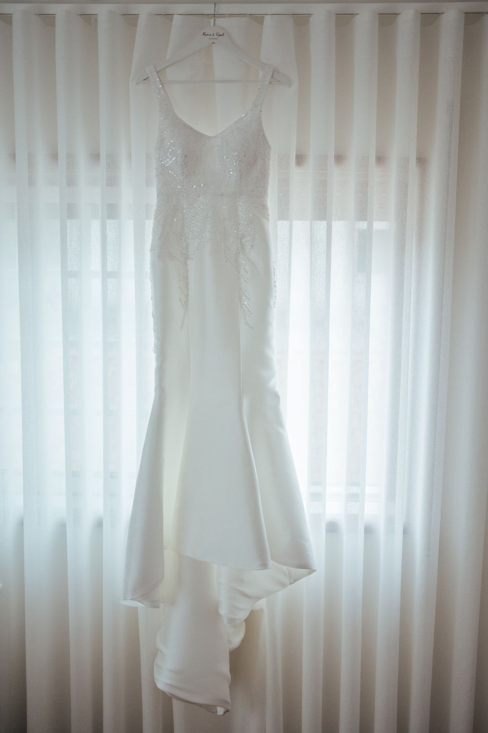 Elegant white mermaid wedding dress with lace bodice and train hanging on a hanger against sheer curtains.