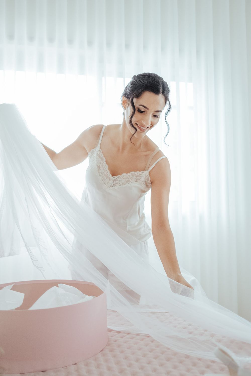 Smiling bride in white lace slip dress arranging her wedding veil on a bed in a bright, airy room.