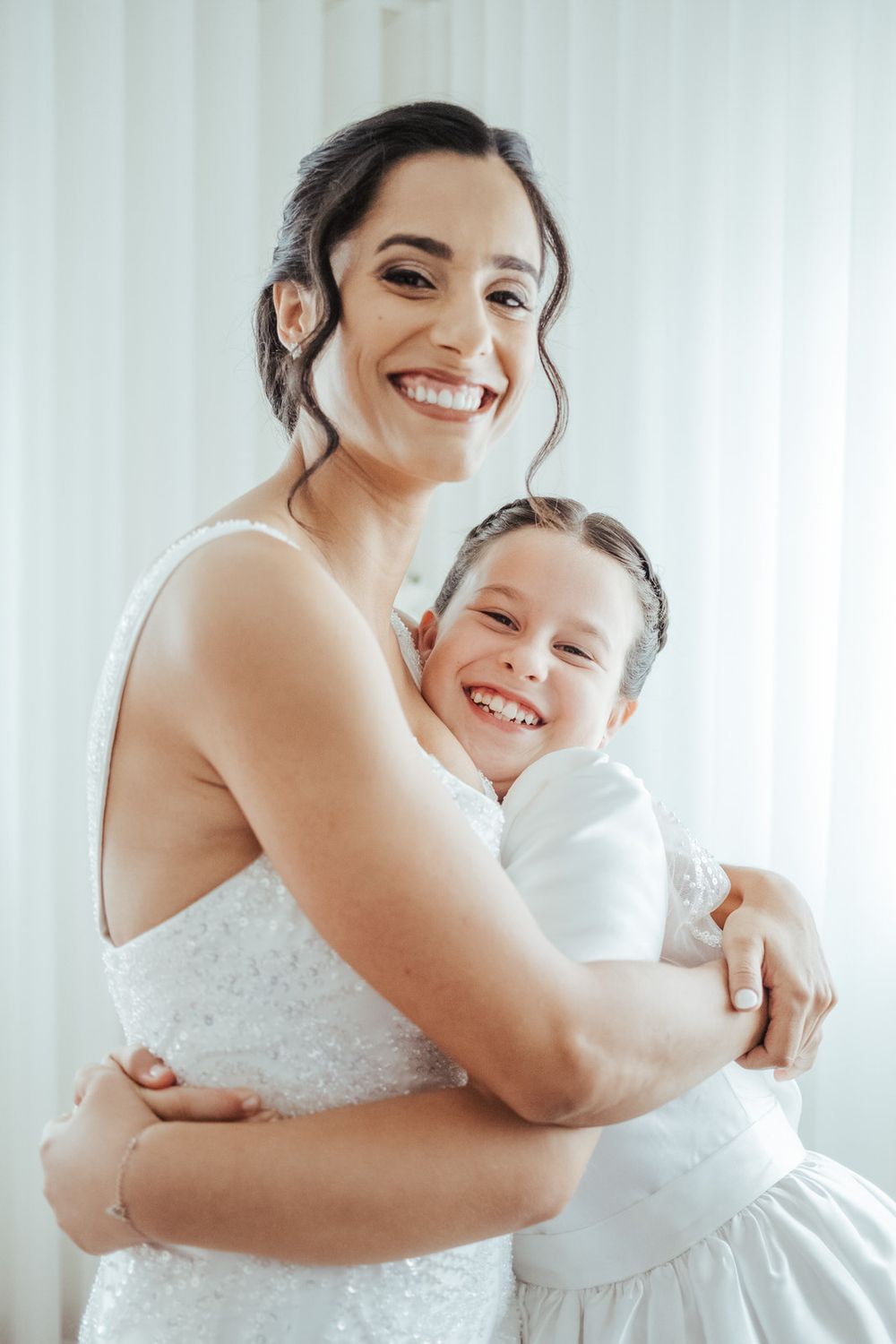 A smiling bride in a white gown hugs a young girl in a white dress, both beaming with joy.