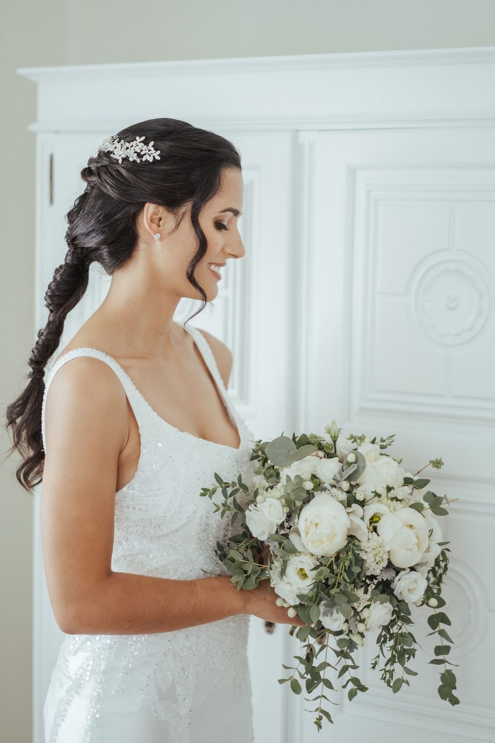 Smiling bride in white gown holding elegant bouquet of white peonies and eucalyptus, with braided hair and floral hair accessory.