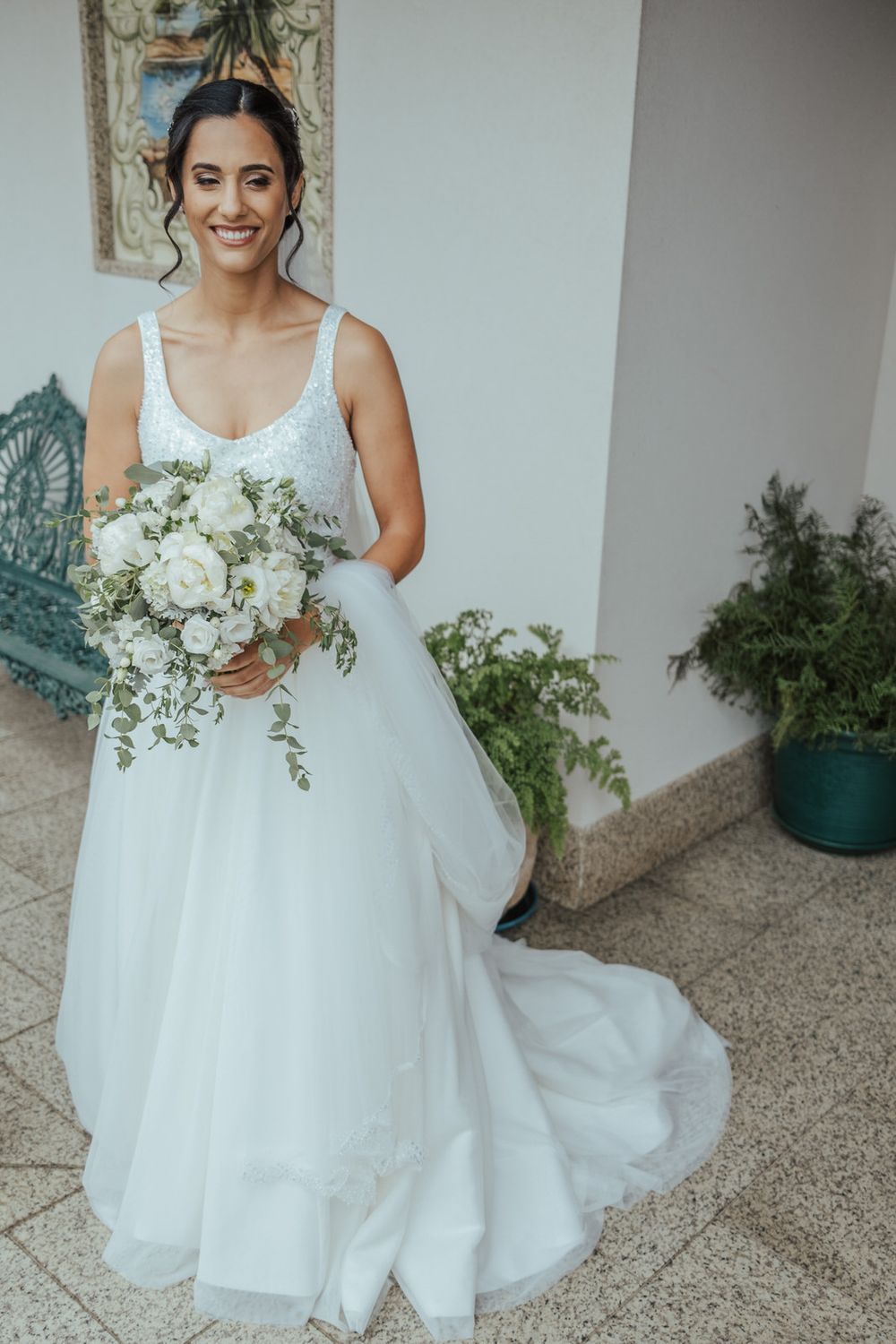 Smiling bride in a white ball gown with beaded bodice holds a white floral bouquet outdoors.