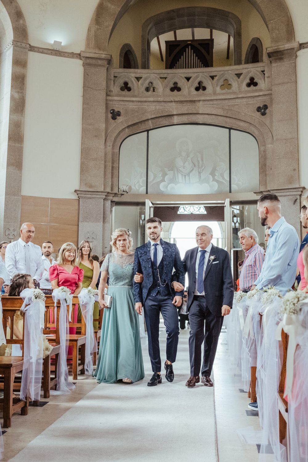 Groom walks down church aisle escorted by parents during elegant wedding ceremony with floral decorations and guests seated in pews.