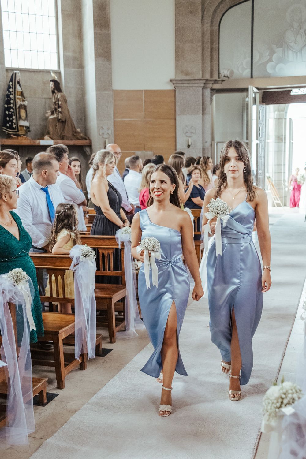 Two bridesmaids in blue satin dresses walk down a church aisle holding white floral bouquets during a wedding ceremony.