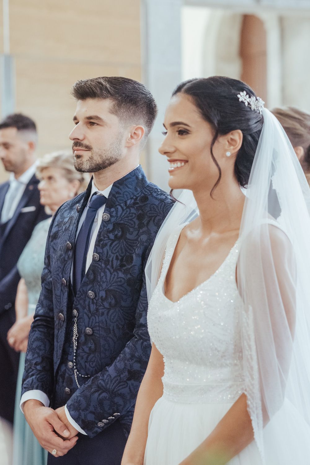 Smiling bride in white gown and veil stands beside groom in navy floral suit during wedding ceremony.