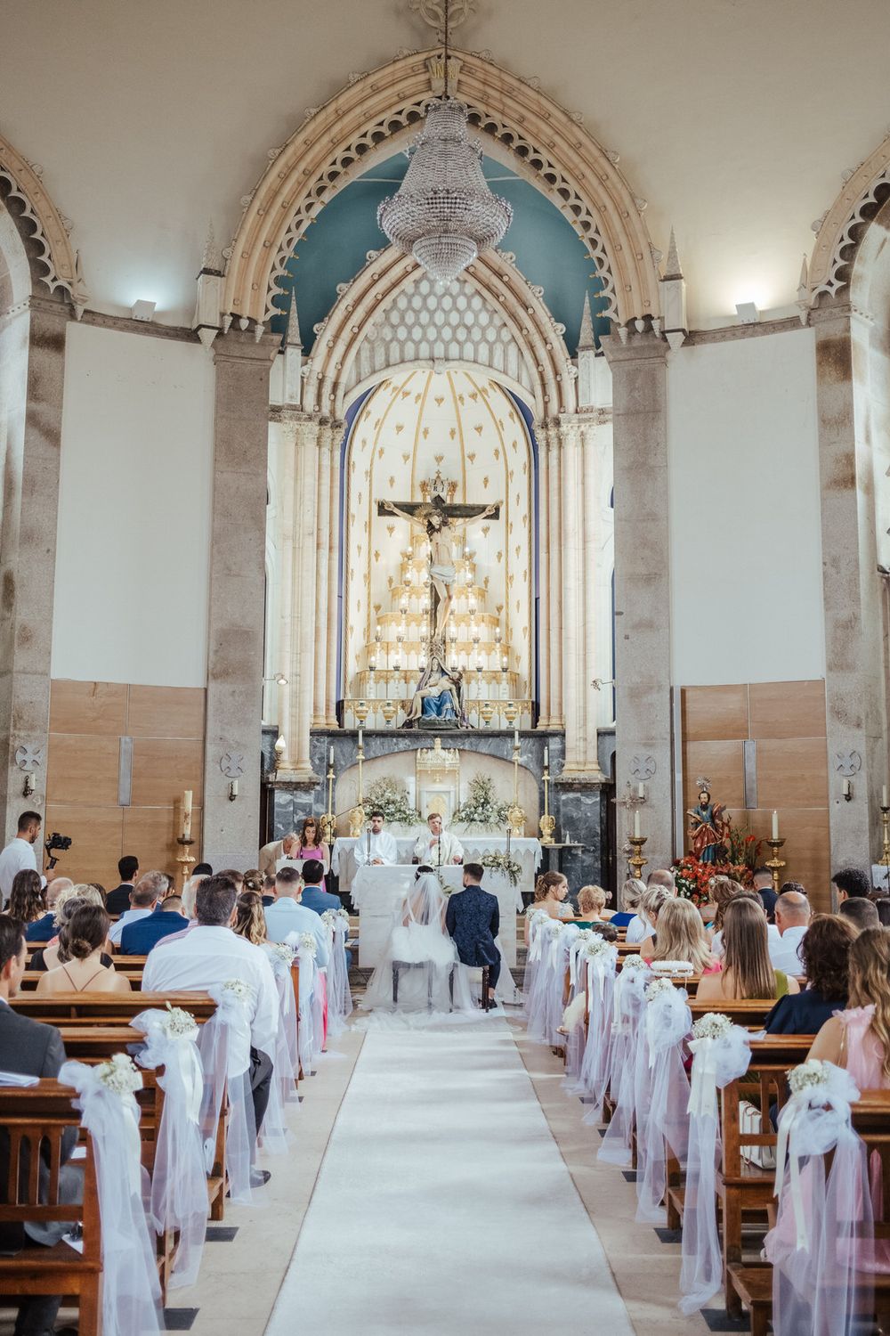 Elegant church wedding ceremony with guests seated in wooden pews along a white aisle leading to a ornate altar.