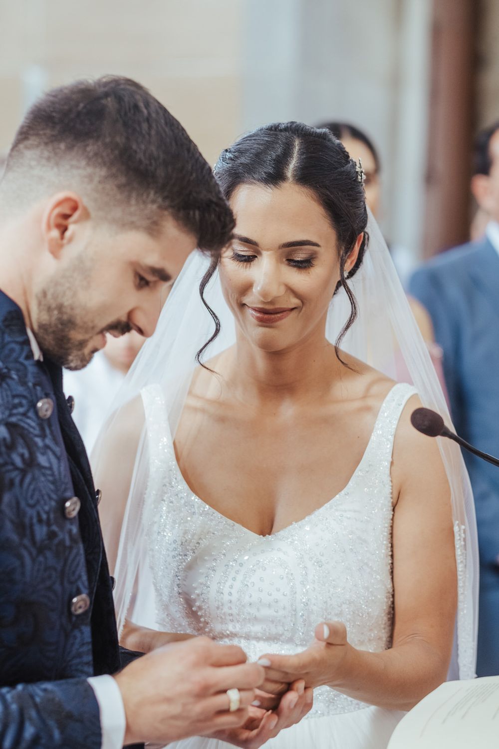 Groom placing wedding ring on brides finger during ceremony, bride smiling in white beaded gown and veil.