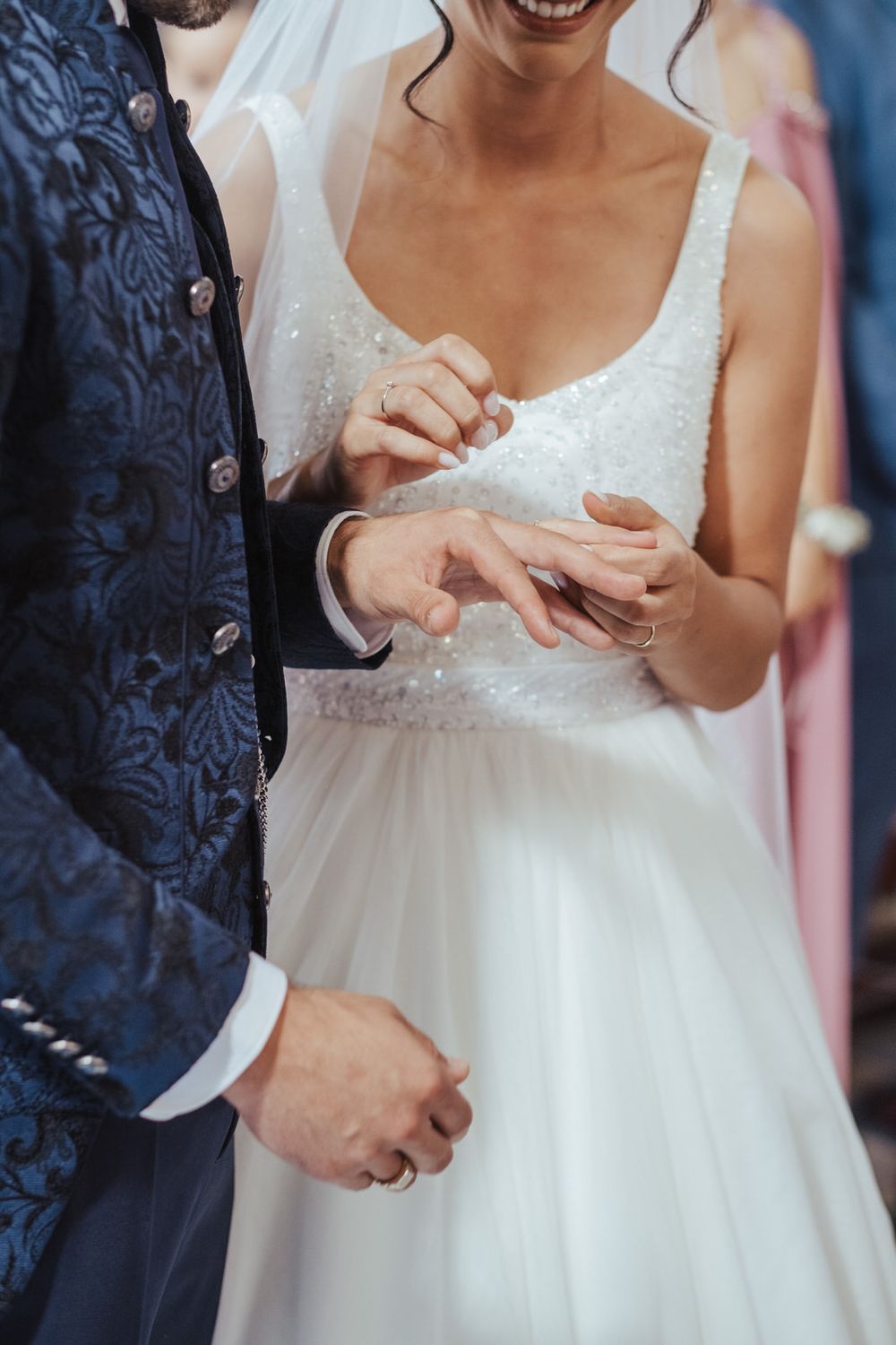 Bride in white gown smiling as she places wedding ring on groom's finger during ceremony.