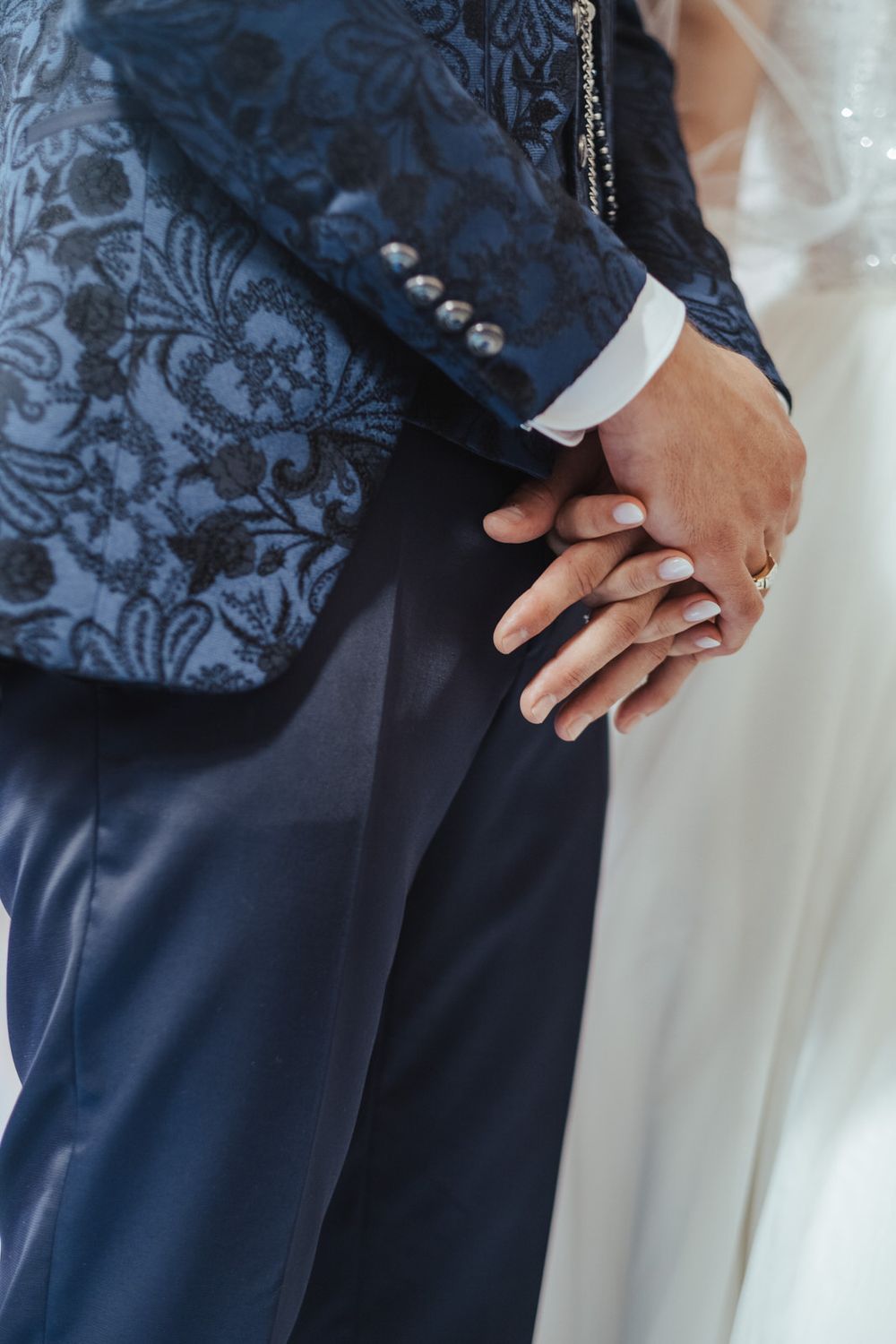 Bride and groom holding hands, groom wearing ornate navy blue floral jacquard suit with wedding rings visible.