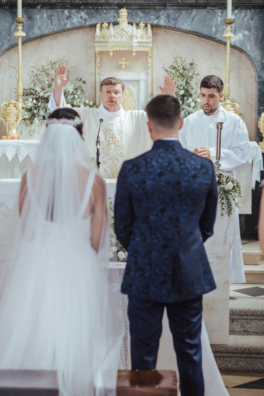 Bride and groom stand before priest at church altar during traditional Catholic wedding ceremony with floral decorations.