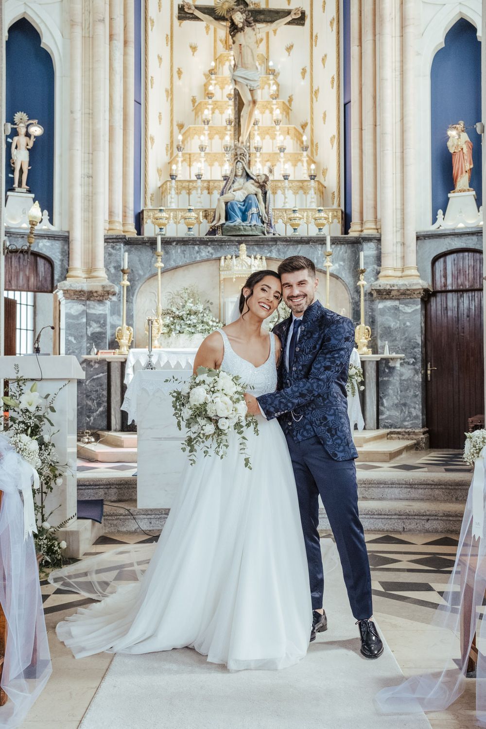 Bride and groom posing together at church altar, bride holding white bouquet, groom in navy floral suit.