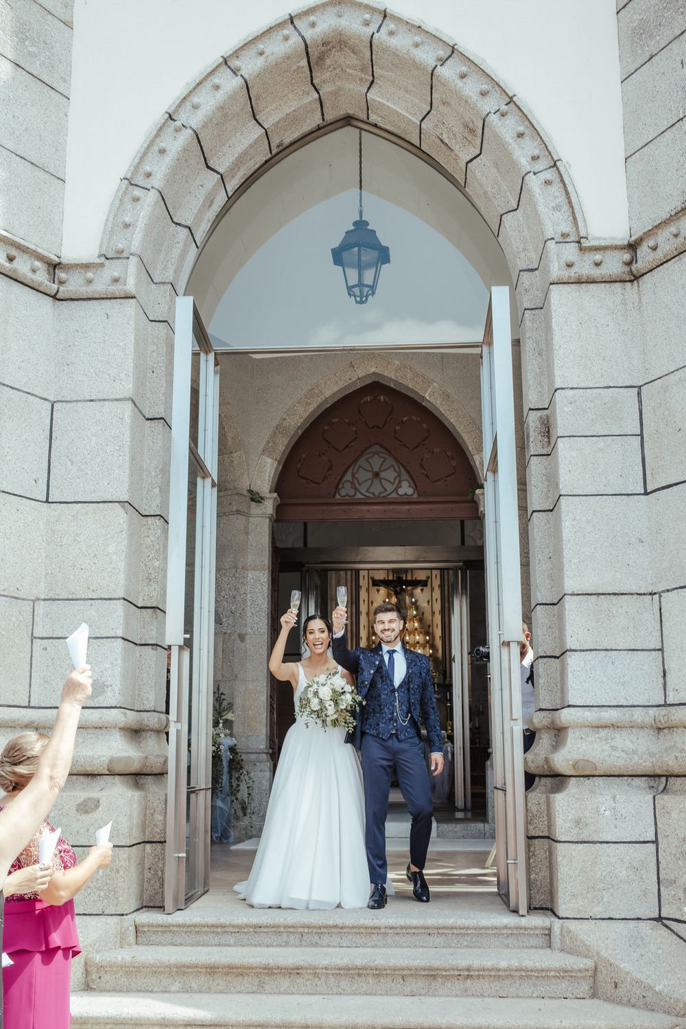 Bride and groom exit stone church doorway, smiling and waving, as guests celebrate on the steps below.