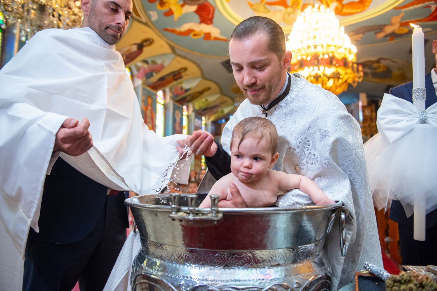 An Orthodox priest baptizes a baby in a silver font during a traditional church ceremony.