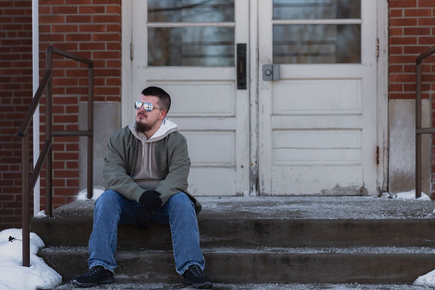 Man in sunglasses sitting on snowy brick building steps in winter, wearing hoodie and jeans.
