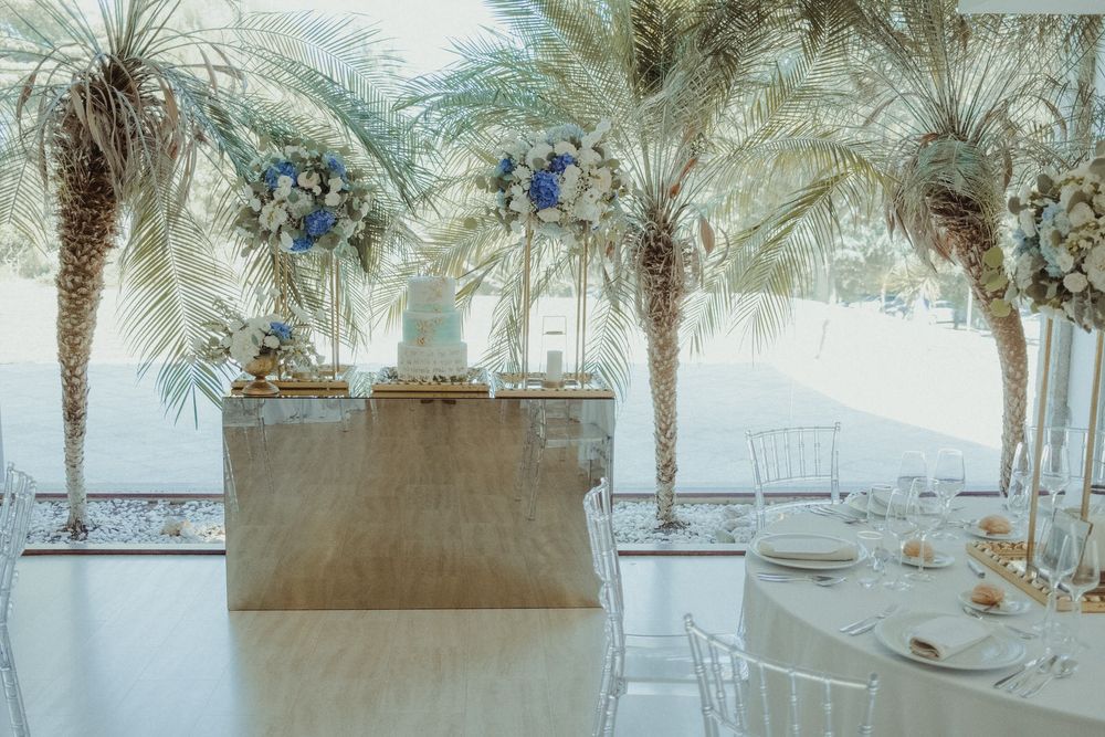 Elegant wedding reception with tall palm trees, blue floral arrangements, and a dessert table in a bright venue.