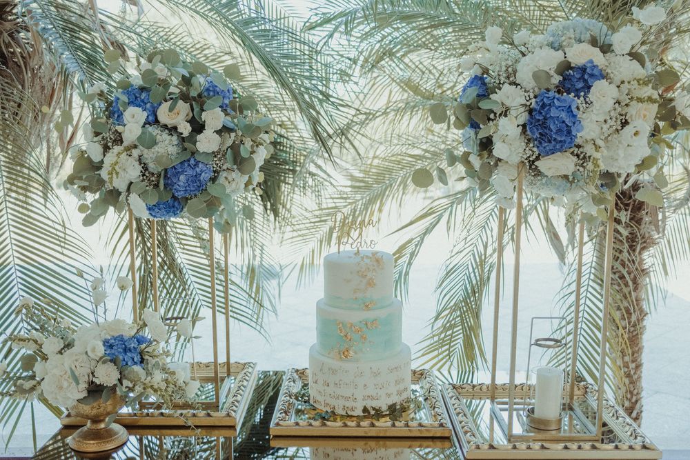 Elegant wedding cake display with blue hydrangeas, white flowers, and gold palm leaf accents on a mirrored table.